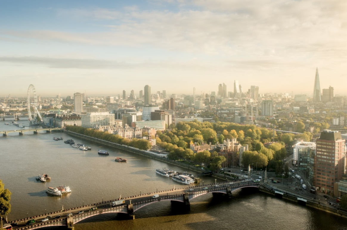 An aerial view of the London skyline featuring the River Thames, a busy bridge, the London Eye on the left, and The Shard in the distant background under a hazy sky.
